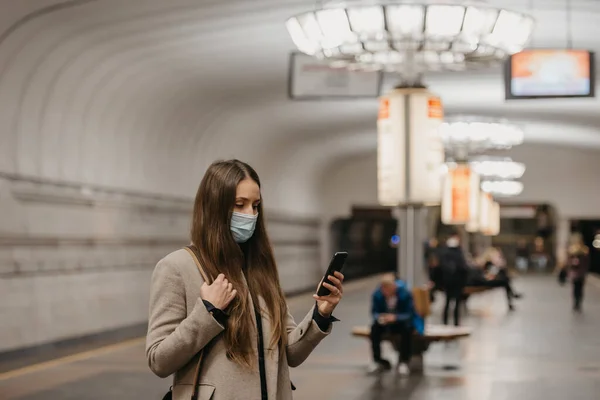 A woman in a face mask to avoid the spread of coronavirus is reading news on a phone at a subway station. A girl in a surgical face mask against COVID-19 is waiting for a train on a metro platform.