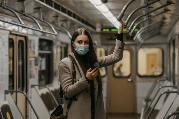 A woman in a medical face mask to avoid the spread of coronavirus is posing in a subway car. A girl with long hair in a surgical mask on her face against COVID-19 is holding a cellphone on a train.