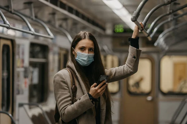 コロナウイルスの拡散を避けるために医療用フェイスマスクの女性が地下鉄の車にポーズをとっています Covid 19に対して彼女の顔に外科用マスクの女の子は電車の中で携帯電話を保持しています — ストック写真