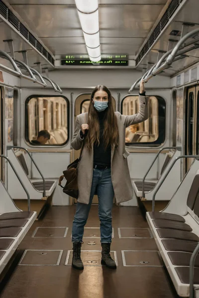 A woman in a medical face mask to avoid the spread of coronavirus is holding onto the handrail in a subway car. A girl with long hair in a surgical mask against COVID-19 is standing on a metro train.