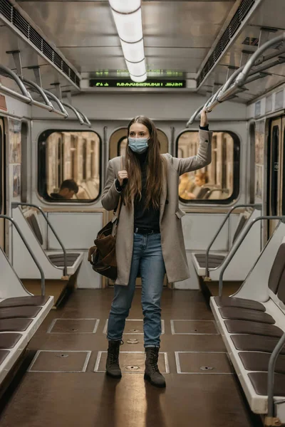 A woman in a face mask to avoid the spread of coronavirus is holding onto the handrail in a subway car. A girl with long hair in a surgical mask against COVID-19 is standing on a metro train.