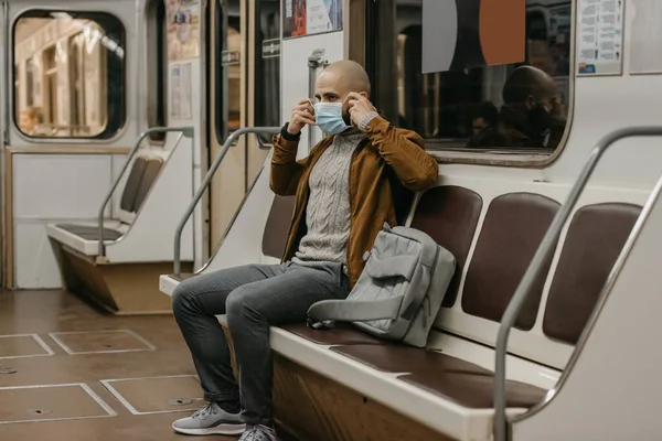 A man with a beard is putting on a medical mask on his face to avoid the spread of coronavirus in a subway car. A bald guy in a surgical face mask against COVID-19 is sitting on a metro train.