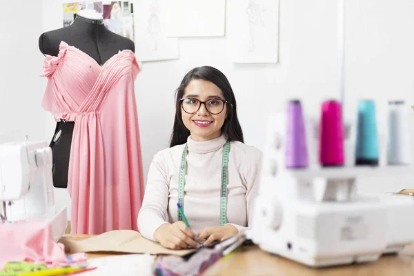 Stüdyoda tasarımları üzerinde çalışan genç bir moda tasarımcısı gülümseyip kameraya bakıyor. Girişimci bir kadın.