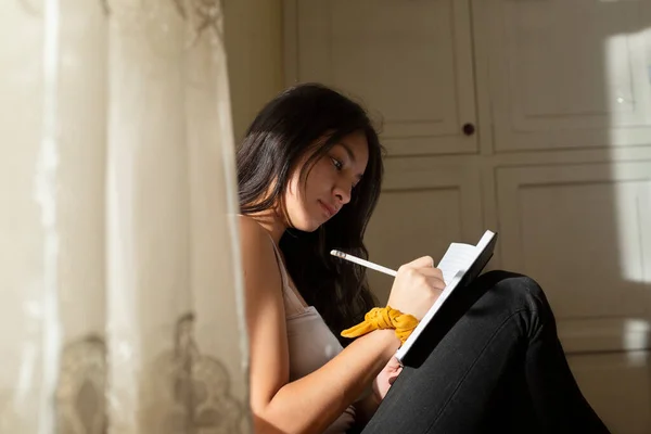 Young hispanic woman writing in a black notebook, thoughtful girl writing in her journal in her room at sunset. young man near the window concentrated studying at home.