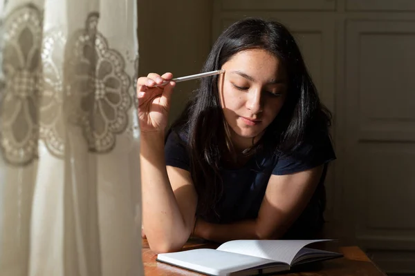 Young Hispanic woman writing in a black notebook, thoughtful girl writing in her journal in her room at sunset. young woman on a cabinet concentrated studying at home.