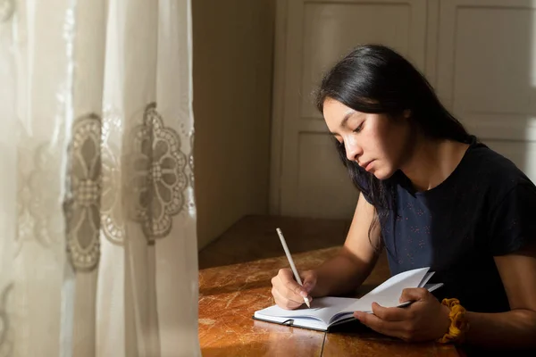 Young Hispanic woman writing in a notebook, serious girl writing in her journal in her room at sunset. concentrated young woman studying at home