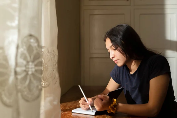 Young Hispanic woman writing in a notebook, serious girl writing in her journal in her room at sunset. concentrated young woman studying at home