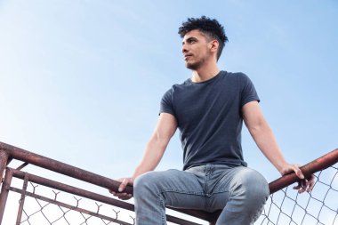 Çatıda oturan İspanyol genç adam günbatımını seyrediyor. Arka planda mavi gökyüzü olan eski metal ağ çerçevesinin tepesindeki adam. Şehir tarzı.