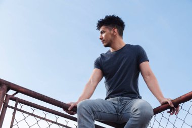 Çatıda oturan İspanyol genç adam günbatımını seyrediyor. Arka planda mavi gökyüzü olan eski metal ağ çerçevesinin tepesindeki adam. Şehir tarzı.
