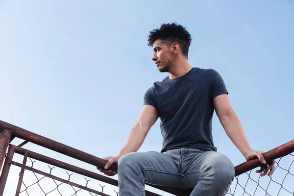 Hispanic young man on rooftop sitting watching sunset - man on top of old metal mesh frame with blue sky in background - urban style