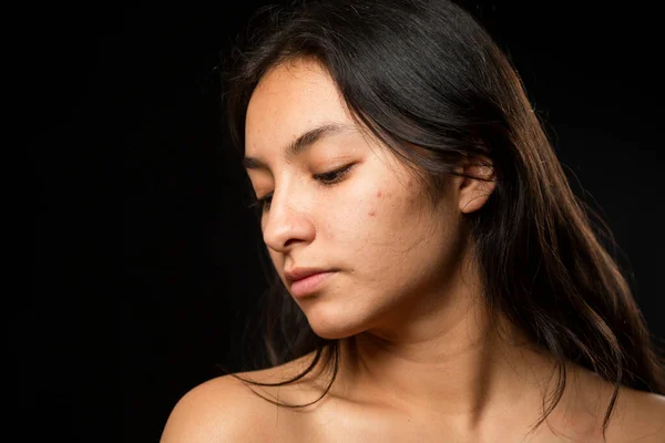 Hispanic woman portrait in studio with black background - Young woman taking care of her natural skin - Beauty portrait, woman looking down unsure of herself
