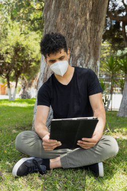 Güneşli bir günde ağaçların altında dijital tabletini kullanan maskeli İspanyol genç adam. Çimenlerde oturan genç adam elektronik aletle çalışıyor. Sosyal uzaklık.