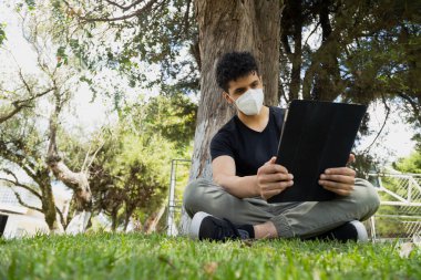 Güneşli bir günde ağaçların altında dijital tabletini kullanan maskeli İspanyol genç adam. Çimenlerde oturan genç adam elektronik aletle çalışıyor. Sosyal uzaklık.