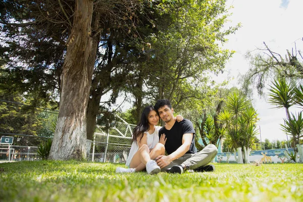 若いヒスパニック系のカップルが草の上に座って楽しんでいます 晴れた日に自然公園を散歩するカップル — ストック写真
