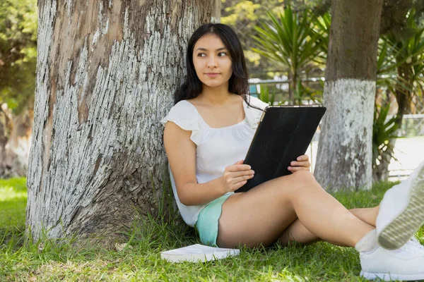 Jovem Hispânico Sentado Sob Uma Árvore Parque Lendo Com Tablet — Fotografia de Stock