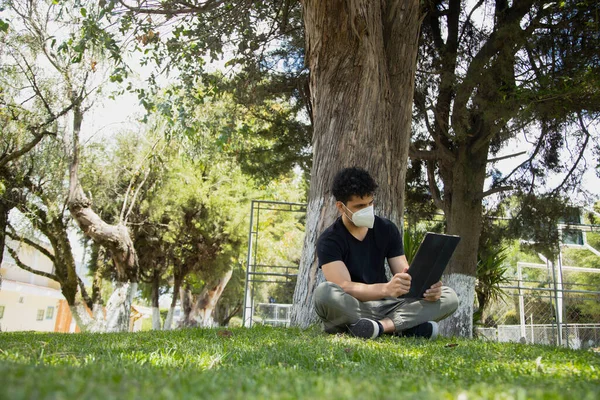 Hispanic young man sitting reading in the park with mask - young man studying with an electronic tablet in a natural park - social distancing - general shot