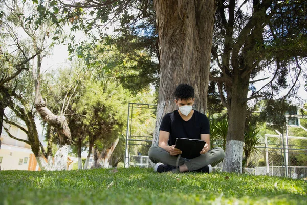 Hispanic young man sitting reading in the park with mask - young man studying with an electronic tablet in a natural park - social distancing