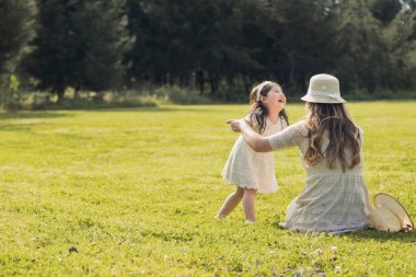 Küçük kızıyla yeşil bir alanda eğlenen genç bir anne. Doğal parkın ortasında beyaz elbiseli bir anne ve kız.