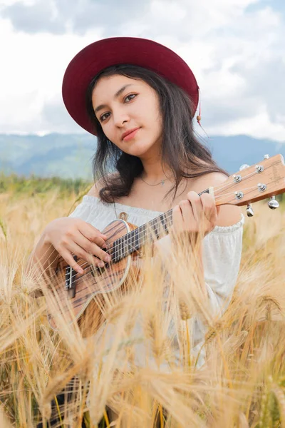 Female artist playing her ukulele in the middle of a golden wheat field - Hipster woman in the middle of a field holding her ukulele - Artist inspired by nature