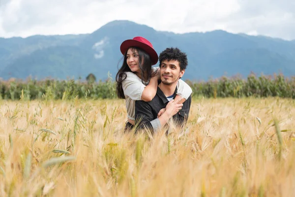 小麦の分野で楽しみを持っている愛のカップル フィールドで一緒に時間を楽しんで幸せな若いヒスパニック系のカップル 夏を楽しんでいる間 男は彼の背中にガールフレンドを運ぶ — ストック写真