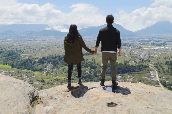 Genç çift dağın tepesinde el ele tutuşup doğanın tadını çıkarıyor. Uçurumun kenarındaki genç insanlar önlerindeki küçük kasabayı izliyor. Quetzaltenango Guatemala 'daki manzaranın tadını çıkarıyorlar..