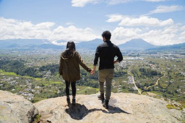 Dağın tepesine doğru yürüyen genç bir çift, bir volkan ve dağlarla çevrili doğanın tadını çıkarıyor. Uçurumun kenarında el ele tutuşmuş genç insanlar, Quetzaltenango Guatemala 'daki açık havanın tadını çıkarıyorlar..