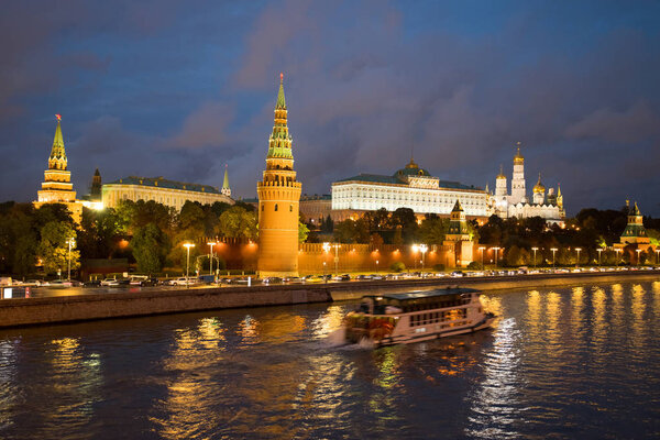 Moscow, Russia. Beautiful View On Moscow Kremlin With Illuminations From Lamps And With Floating Motor Ship On Coast Of Moscow River Under Blue Dramatic Sky In Evening Day In Autumn.