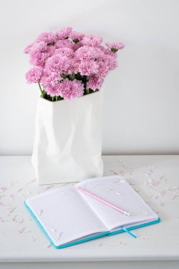 Güzel natürmort defter kalem ve yakınındaki beyaz vazo pembe yaprakları çiçeklerle beyaz masaya çiçek ile kafeste.