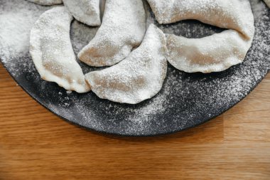 Koyu tabakta ev yapımı hamur tatlısı ve ahşap masa üzerinde un. Ukraynalı vareniki. Kapatın. Üst görünüm.