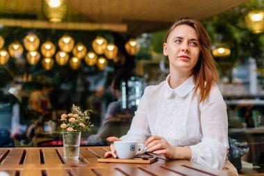 Kafede bir fincan kahve tutan genç ve güzel bir kadın. Tatilde seyahat ederken poz veren kadın blog yazarının açık hava portresi. Yeni fikirleri düşünen serbest çalışan mutlu insanlar. Uzaklara bakıyorum.