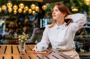 Kafede bir fincan kahve tutan genç ve güzel bir kadın. Tatilde seyahat ederken poz veren kadın blog yazarının açık hava portresi. Yeni fikirleri düşünen serbest çalışan mutlu insanlar. Uzaklara bakıyorum.