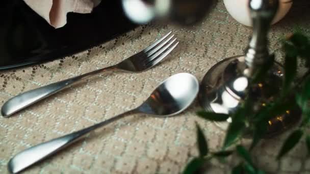 Nahaufnahme eines Löffels und einer Gabel, die auf einer Tischdecke rechts neben einem schwarzen Teller liegen und durch einen Kerzenständer schießen — Stockvideo
