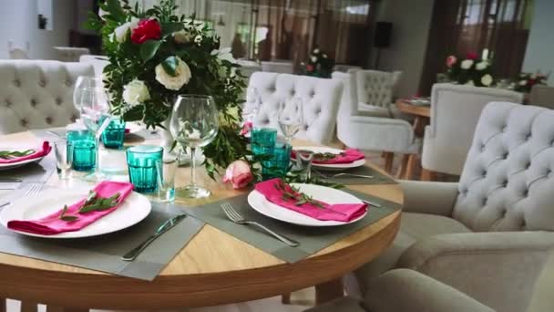 Een ronde houten tafel versierd met bloemstukken gemaakt van witte platen met roze servetten rond die er zachte stoelen zijn — Stockvideo