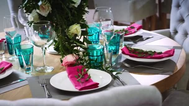 Ein runder Holztisch mit floralen Arrangements aus weißen Tellern mit rosa Servietten, um die herum weiche Stühle stehen — Stockvideo