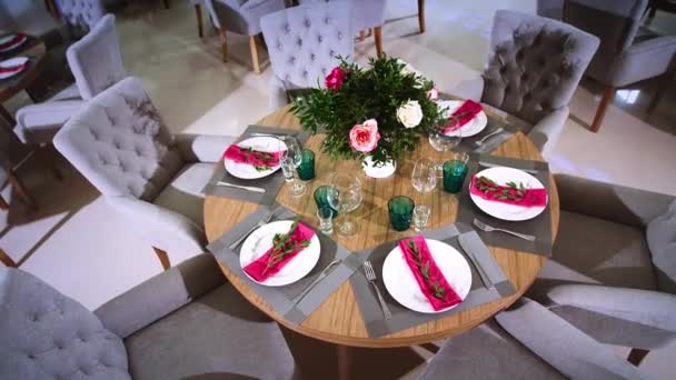 Ένα στρογγυλό ξύλινο τραπέζι διακοσμημένο με floral ρυθμίσεις από λευκές πλάκες με ροζ χαρτοπετσέτες γύρω από την οποία υπάρχουν μαλακά καρέκλες — Αρχείο Βίντεο