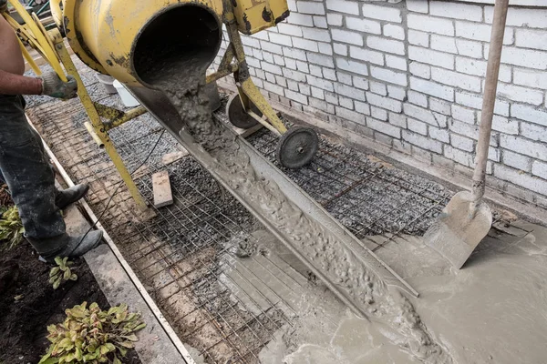 Construction of a concrete walkway around a residential rural house in manual mode. Manually cooked mortar in a concrete mixer. Leveling the surface of the pavement with a special tool