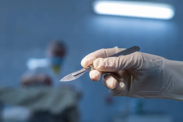 Surgical steel simple shiny scalpel in the surgeon\'s hand against the background of the operation. The faces of the people in the background are unrecognizable in full blur. Conceptual photography. Symbol of surgery,  doctor