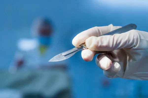 Surgical steel simple shiny scalpel in the surgeon\'s hand against the background of the operation. The faces of the people in the background are unrecognizable in full blur. Conceptual photography. Symbol of surgery,  doctor