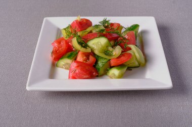 Beyaz kare parlak bir tabakta salatalık, domates ve biber örneği. Sağlıklı beslenme ve vejetaryenliğin sembolü olarak bez bir masa örtüsü üzerine servis tabağı.