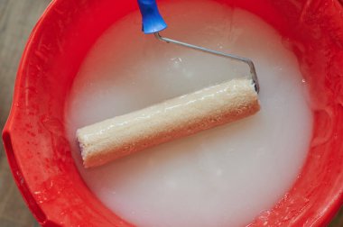 Duvarlara uygulanmadan önce duvar kağıdına yapıştırıcı sürmekte kullanılan bir boya silindiri. Örgü beyaz, kap parlak kırmızı, ressamın aleti iş başında.