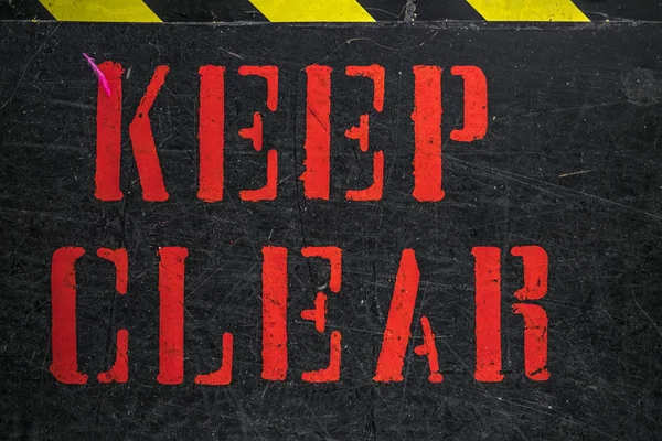 Keep Clear sign painted with capital red letters on scratched black surface, viewed in closeup full frame. Black and yellow attention stripe on the top of the frame