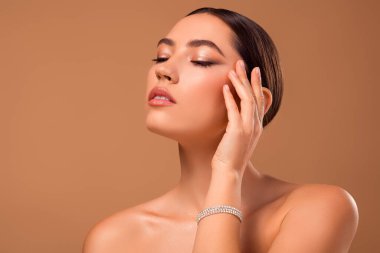 Bej bir arka plana karşı poz veren dingin genç bir kadın kusursuz cildini, şık makyajını ve şık görünümünü özgüven ve zarafet havasıyla entelektüelliği ve zarafeti yansıtıyor..