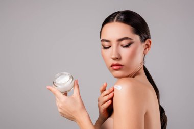 Genç, güzel bir kadın elinde bir kavanozla cildine krem sürüyor, pürüzsüz cilt bakımı uygulamalarını ve minimalist arka plandaki güzelliği vurguluyor..