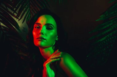 Tropikal yapraklarla çevrili, çarpıcı yeşil ve kırmızı ışıklandırmayla çarpıcı bir şekilde aydınlatılmış güzel bir genç kadının zarif portresi.