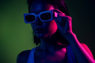 Neon ışıkta poz veren, şık güneş gözlüğü takan genç bir kadın. Renkli arkaplan onun modern ve büyüleyici tarzını geliştirir, canlı ve modaya uygun bir atmosfer yakalar..