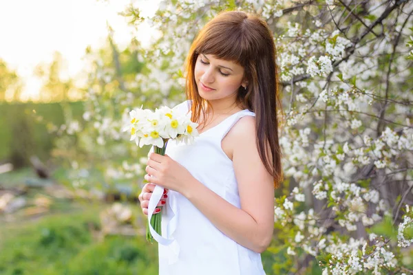 Schöne junge Mädchen mit einem Strauß Narzissen in einem weißen Kleid auf einem Hintergrund von Kirschblüten — Stockfoto