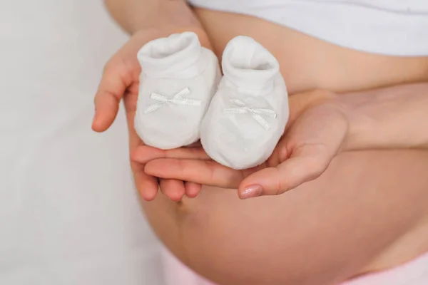 Chaussons pour nouveau-né dans les mains d'une fille enceinte sur le fond de l'abdomen — Photo