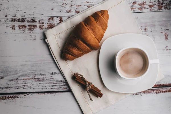 Weiße Tasse mit Kaffee und Croissant und Zimt auf Holztisch. Traditionelles französisches Frühstück am Morgen — Stockfoto