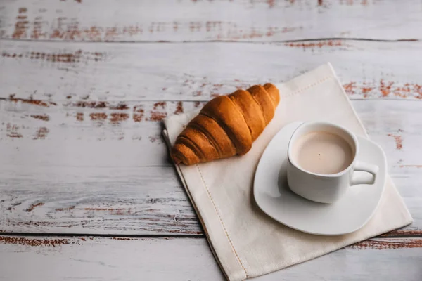 Croissant und eine weiße Tasse Kaffee auf einem Holztisch — Stockfoto