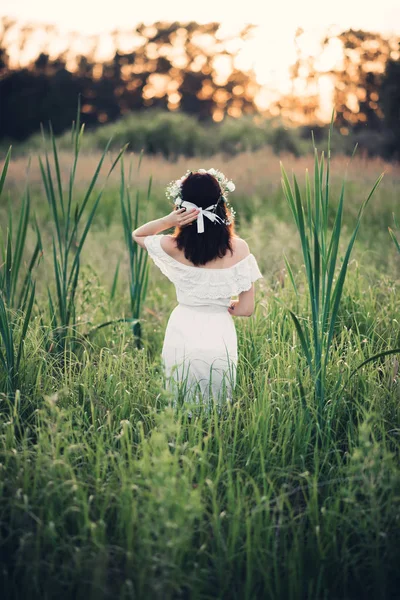 Dívka v bílých šatech s květinovým věncem stojí v poli v létě — Stock fotografie